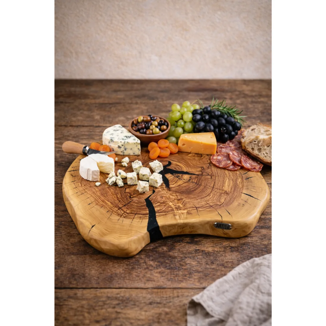 Tábua artesanal de madeira de oliveira em rodela natural, com detalhe preto, usada como tábua de servir com queijos, enchidos, pão, uvas, azeitonas e frutos secos, sobre uma mesa rústica.